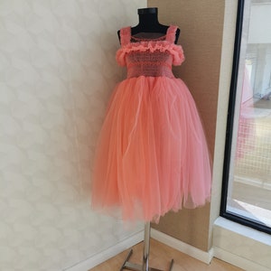 K&ouml;nnte beinhalten: Korallfarbenes T&uuml;llkleid auf einer Schaufensterpuppe. Das Oberteil hat ein detailliertes, strukturiertes Design mit quadratischem Ausschnitt und Tr&auml;gern. Der volle, geschichtete Rock erzeugt eine volumin&ouml;se Silhouette. Das Kleid steht auf einem silbernen Metallst&auml;nder.