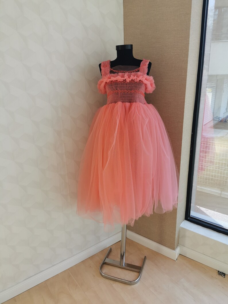 K&ouml;nnte beinhalten: Korallfarbenes T&uuml;llkleid auf einer Schaufensterpuppe. Das Oberteil hat ein detailliertes, strukturiertes Design mit quadratischem Ausschnitt und Tr&auml;gern. Der volle, geschichtete Rock erzeugt eine volumin&ouml;se Silhouette. Das Kleid steht auf einem silbernen Metallst&auml;nder.