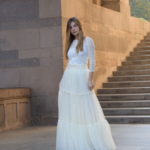 May include: A white, tiered maxi skirt and a white, long-sleeved top. The top has a textured, grid-like pattern. The skirt is made of a flowing, lightweight fabric. The outfit is suitable for a wedding or special occasion.