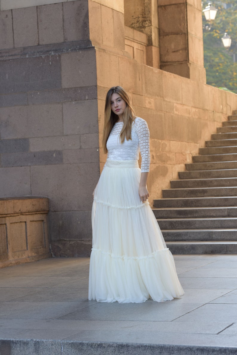 May include: A white, tiered maxi skirt and a white, long-sleeved top. The top has a textured, grid-like pattern. The skirt is made of a flowing, lightweight fabric. The outfit is suitable for a wedding or special occasion.