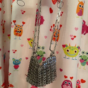 May include: A silver chainmail shoulder bag with a long chain strap. The bag is made of small, interlocking metal rings, creating a textured, draped effect. The background features a pink fabric with cartoon monsters and hearts.