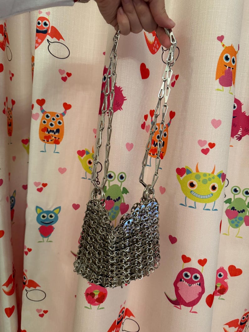 May include: A silver chainmail shoulder bag with a long chain strap. The bag is made of small, interlocking metal rings, creating a textured, draped effect. The background features a pink fabric with cartoon monsters and hearts.