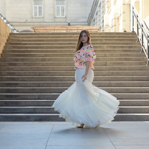 May include: A woman wearing a white, tiered, floor-length gown and a colorful top with ruffles. The top has a pink base with neon accents. The background features a large staircase and a building.