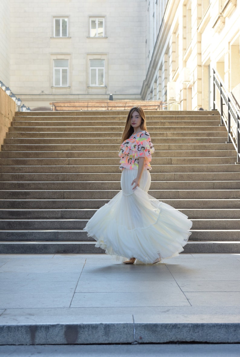 May include: A woman wearing a white, tiered, floor-length gown and a colorful top with ruffles. The top has a pink base with neon accents. The background features a large staircase and a building.