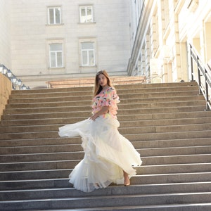 May include: A woman wearing a layered white skirt and a top with colorful accents stands on a wide stone staircase. The skirt flows as she moves, creating a dynamic image. The background features a large building with multiple windows.
