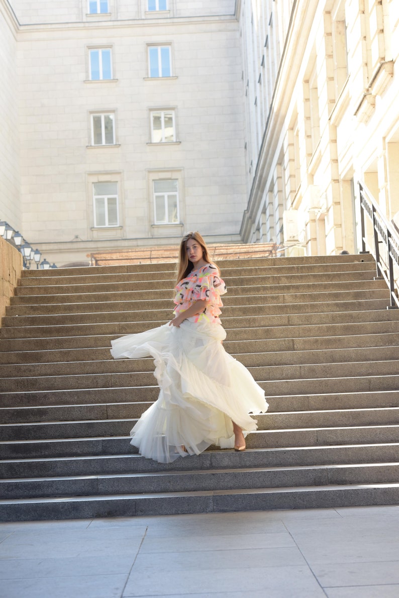 May include: A woman wearing a layered white skirt and a top with colorful accents stands on a wide stone staircase. The skirt flows as she moves, creating a dynamic image. The background features a large building with multiple windows.