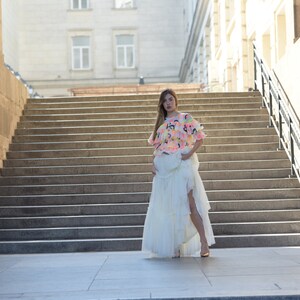 May include: A woman wearing a colorful top and a long white skirt stands on a set of outdoor stairs. The top is a mix of pink, yellow, and blue, while the skirt has a layered design with a high slit.