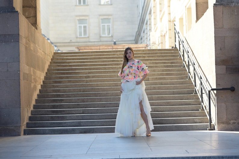 May include: A woman wearing a colorful top and a long white skirt stands on a set of outdoor stairs. The top is a mix of pink, yellow, and blue, while the skirt has a layered design with a high slit.
