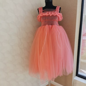 K&ouml;nnte beinhalten: Korallfarbenes Kleid auf einer schwarzen Schaufensterpuppe. Das Kleid hat einen T&uuml;llrock und ein Oberteil mit R&uuml;schentr&auml;gern und Details. Das Kleid wird auf einem silbernen St&auml;nder vor einer neutralen Wand pr&auml;sentiert.