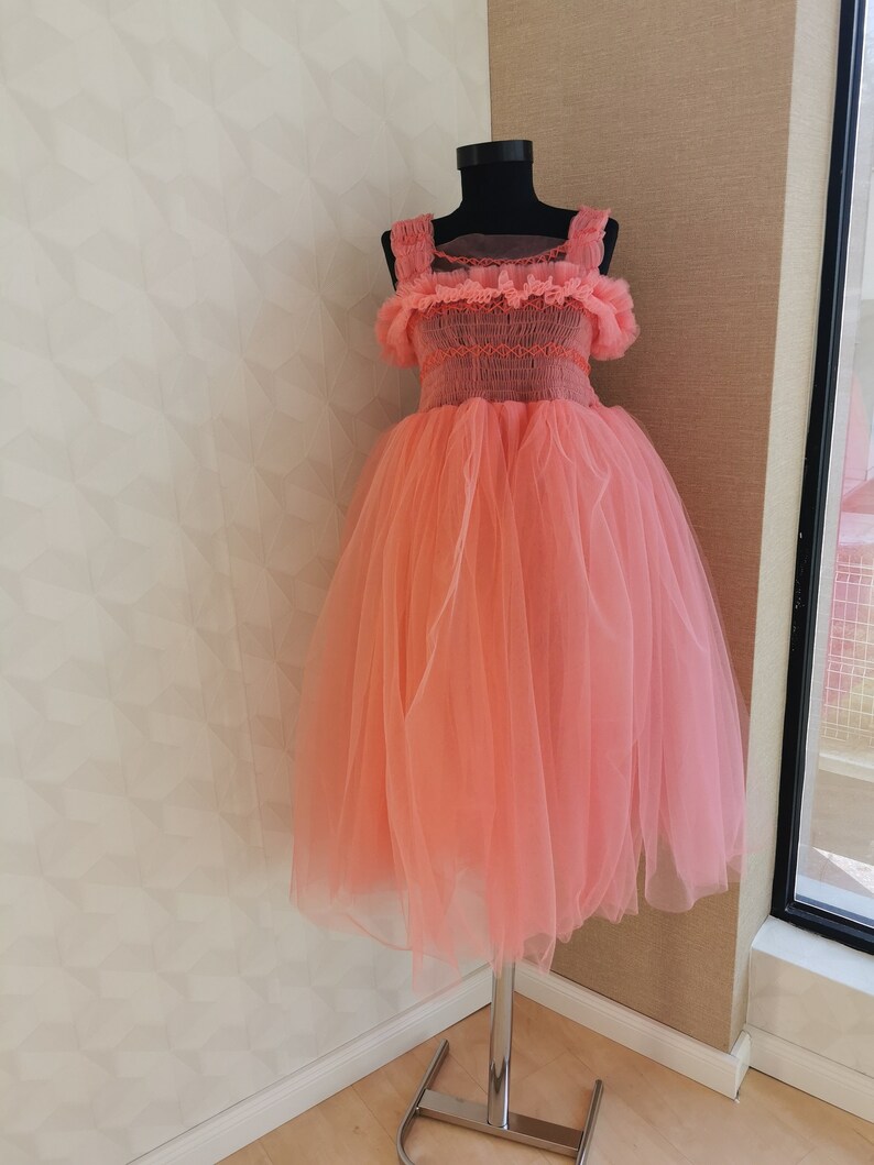 K&ouml;nnte beinhalten: Korallfarbenes Kleid auf einer schwarzen Schaufensterpuppe. Das Kleid hat einen T&uuml;llrock und ein Oberteil mit R&uuml;schentr&auml;gern und Details. Das Kleid wird auf einem silbernen St&auml;nder vor einer neutralen Wand pr&auml;sentiert.