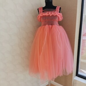 K&ouml;nnte beinhalten: Korallfarbenes Kleid auf einer schwarzen Schaufensterpuppe. Das Kleid hat einen T&uuml;llrock und ein Oberteil mit R&uuml;schentr&auml;gern und Details. Das Kleid wird auf einem silbernen St&auml;nder vor einer neutralen Wand pr&auml;sentiert.