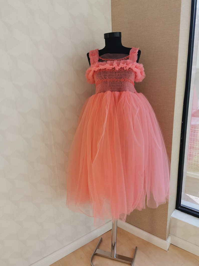 K&ouml;nnte beinhalten: Korallfarbenes Kleid auf einer schwarzen Schaufensterpuppe. Das Kleid hat einen T&uuml;llrock und ein Oberteil mit R&uuml;schentr&auml;gern und Details. Das Kleid wird auf einem silbernen St&auml;nder vor einer neutralen Wand pr&auml;sentiert.