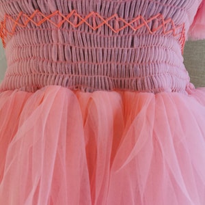 K&ouml;nnte beinhalten: Ein rosa Kleid mit einem Oberteil mit Smok-Design und orangefarbenen Kreuzstichdetails. Der Rock besteht aus Lagen aus weichem, transparentem rosa T&uuml;ll. Das Kleid hat eine romantische, luftige &Auml;sthetik.