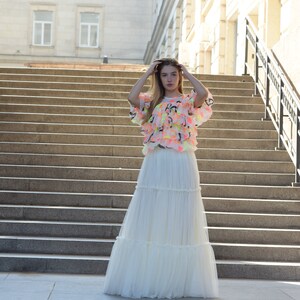 May include: A woman wearing a tiered white maxi skirt and a ruffled top with neon accents. The top has a light pink base with yellow, green, and black details. The outfit is worn outdoors in front of a large staircase.