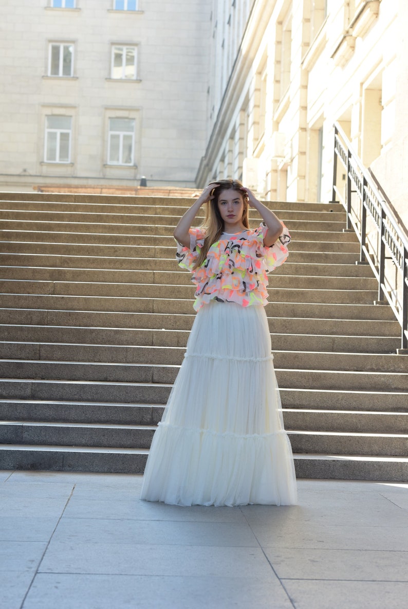 May include: A woman wearing a tiered white maxi skirt and a ruffled top with neon accents. The top has a light pink base with yellow, green, and black details. The outfit is worn outdoors in front of a large staircase.
