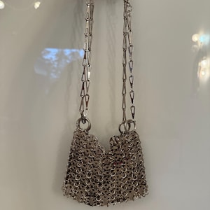 May include: A silver chainmail bag with a shoulder strap made of linked metal pieces. The bag's body is constructed from small, interlocking metal rings, creating a textured, shimmering surface. The bag is suspended by a chain strap.