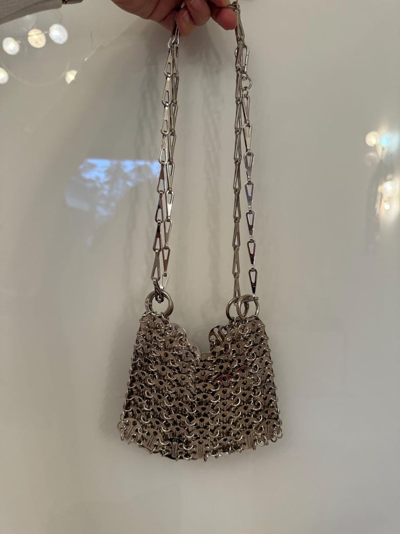 May include: A silver chainmail bag with a shoulder strap made of linked metal pieces. The bag's body is constructed from small, interlocking metal rings, creating a textured, shimmering surface. The bag is suspended by a chain strap.