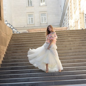 May include: A woman in a tiered white tulle skirt and a colorful top stands on a stone staircase. The top features pink, yellow, and orange accents. The background includes a large building with multiple windows.