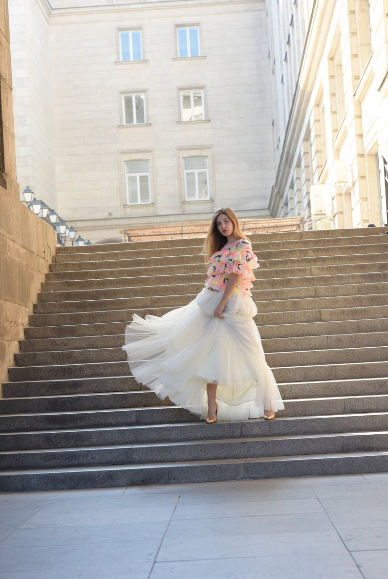 May include: A woman in a tiered white tulle skirt and a colorful top stands on a stone staircase. The top features pink, yellow, and orange accents. The background includes a large building with multiple windows.