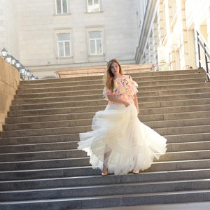 May include: A woman in a flowing white dress and a colorful top stands on a grand staircase. The dress has multiple layers, and the top features a vibrant pattern of pink, yellow, and blue. Gold heels complete the look.