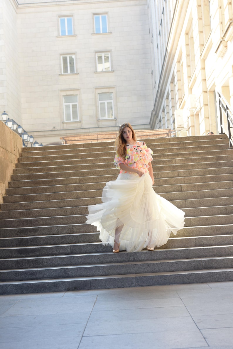 May include: A woman in a flowing white dress and a colorful top stands on a grand staircase. The dress has multiple layers, and the top features a vibrant pattern of pink, yellow, and blue. Gold heels complete the look.