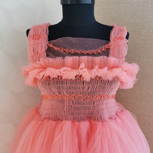 K&ouml;nnte beinhalten: Korallfarbenes Kleid auf einer schwarzen Schaufensterpuppe. Das Kleid hat ein gerafftes Oberteil mit orangefarbenen N&auml;hten, R&uuml;schenbesatz und einen mehrlagigen T&uuml;llrock. Die Tr&auml;ger sind ebenfalls ger&uuml;scht, und das Kleid hat einen transparenten &Uuml;berzug.