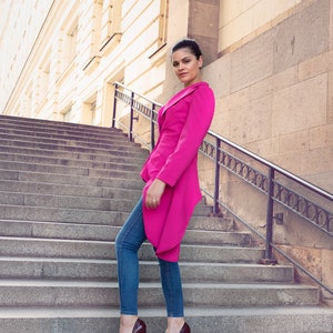 May include: A woman wearing a bright pink blazer, blue jeans, and burgundy high heels stands on a set of stone steps. The blazer has a unique asymmetrical design. The background features a building with a stone facade.