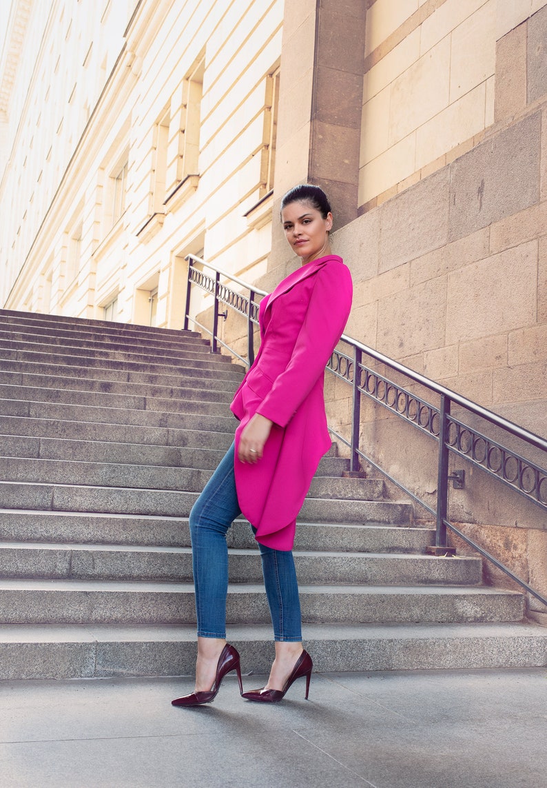 May include: A woman wearing a bright pink blazer, blue jeans, and burgundy high heels stands on a set of stone steps. The blazer has a unique asymmetrical design. The background features a building with a stone facade.