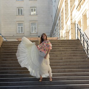 May include: A woman wearing a white tulle skirt and a colorful top with a pink, yellow, and black design. The woman is standing on a set of stone steps in front of a building.
