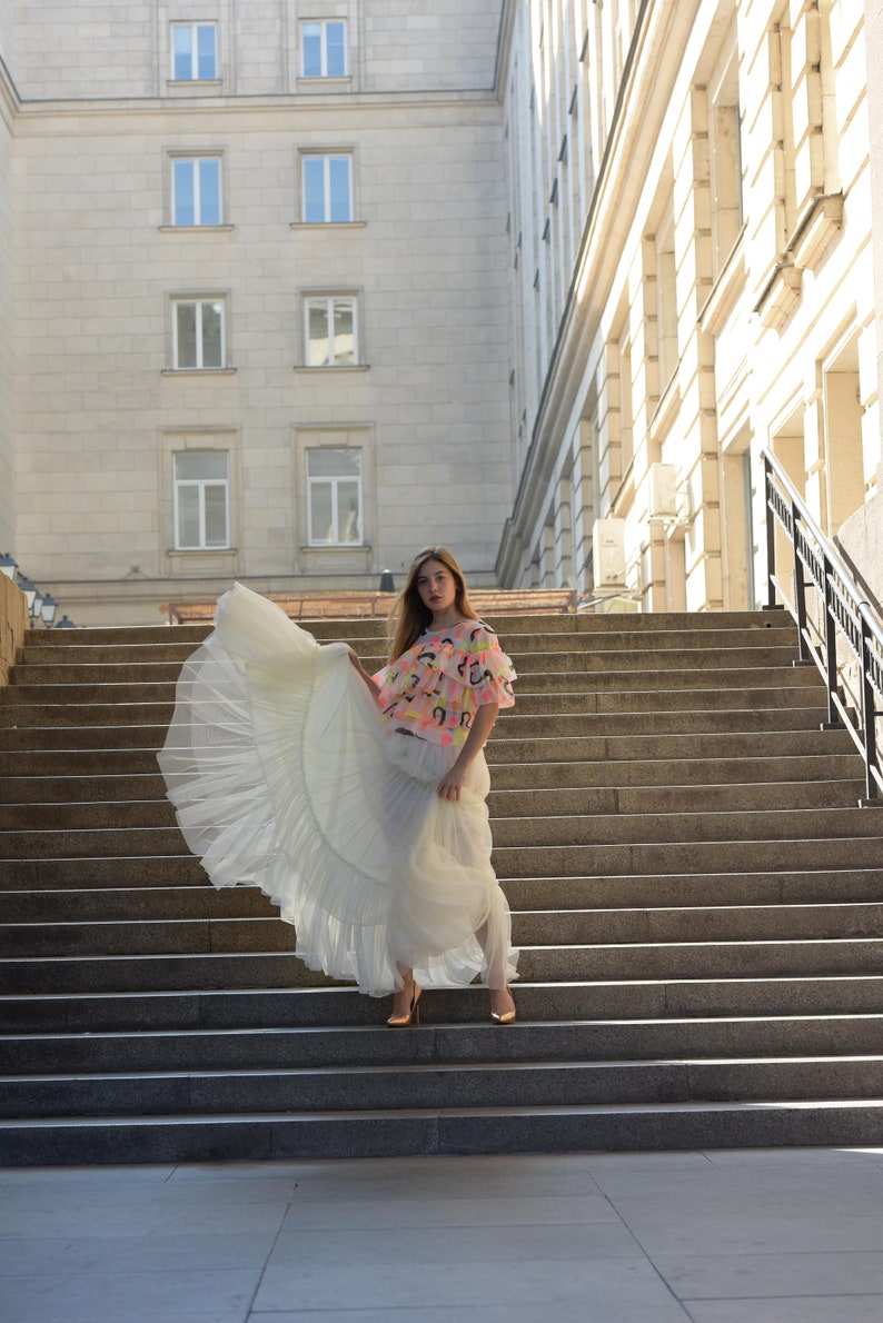 May include: A woman wearing a white tulle skirt and a colorful top with a pink, yellow, and black design. The woman is standing on a set of stone steps in front of a building.