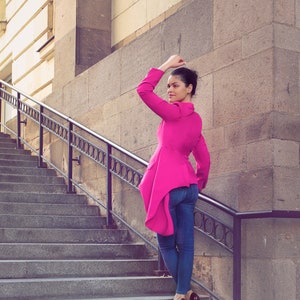 May include: A woman wearing a bright pink jacket and blue jeans stands on a stone staircase. The jacket has a unique asymmetrical design. She is wearing dark high heels. The background features a stone building and a metal railing.