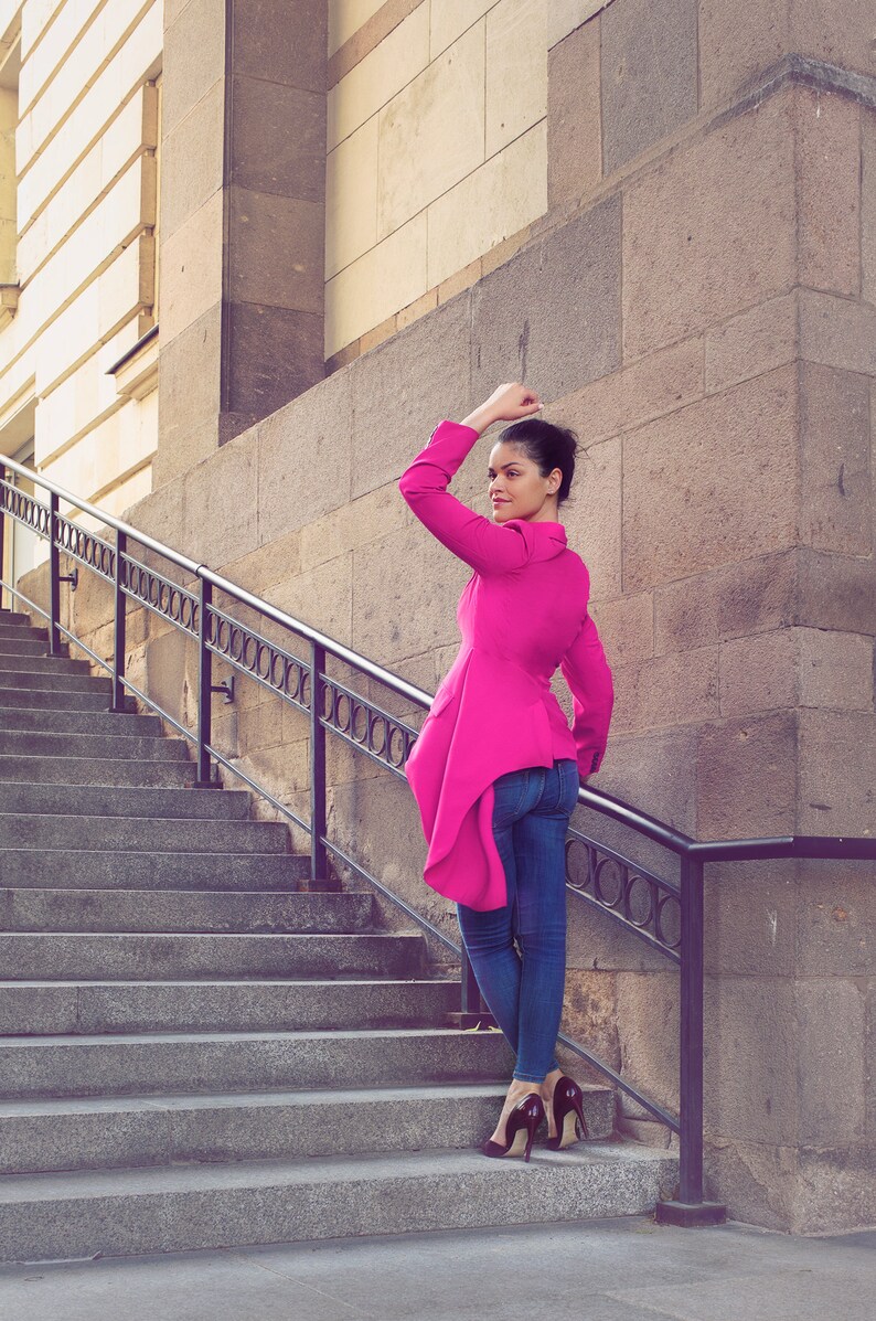 May include: A woman wearing a bright pink jacket and blue jeans stands on a stone staircase. The jacket has a unique asymmetrical design. She is wearing dark high heels. The background features a stone building and a metal railing.
