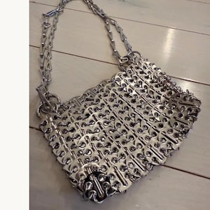 May include: A silver handbag crafted from recycled can tabs. It features a chain strap and a diamond-shaped body. The can tabs are interlinked, forming a textured, metallic surface. The bag is displayed on a light-coloured wooden surface.