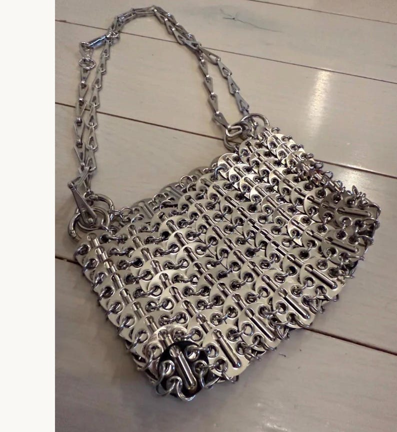 May include: A silver handbag crafted from recycled can tabs. It features a chain strap and a diamond-shaped body. The can tabs are interlinked, forming a textured, metallic surface. The bag is displayed on a light-coloured wooden surface.