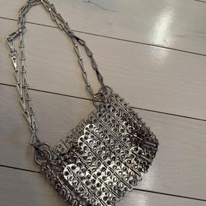 May include: A silver handbag with a chain strap. The bag is made from interlocking metal pieces, forming a textured, geometric design. The silver chain strap attaches to the bag with metal rings. The bag has a rectangular shape.