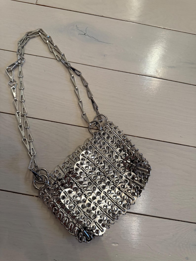 May include: A silver handbag with a chain strap. The bag is made from interlocking metal pieces, forming a textured, geometric design. The silver chain strap attaches to the bag with metal rings. The bag has a rectangular shape.