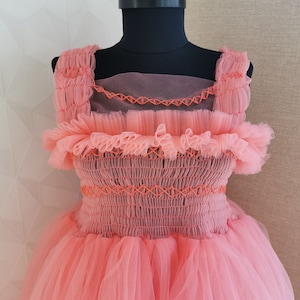 K&ouml;nnte beinhalten: Korallfarbenes Kleid auf einer schwarzen Schaufensterpuppe. Das Kleid hat ein gerafftes Oberteil mit orangefarbenen N&auml;hten, R&uuml;schenbesatz und einen mehrlagigen T&uuml;llrock. Die Tr&auml;ger sind ebenfalls ger&uuml;scht, und das Kleid hat einen transparenten &Uuml;berzug.