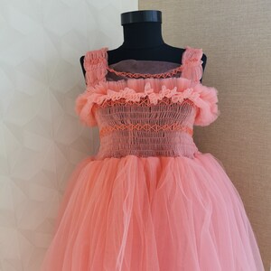 K&ouml;nnte beinhalten: Ein pfirsichfarbenes T&uuml;llkleid, das auf einer schwarzen Schaufensterpuppe pr&auml;sentiert wird. Das Kleid hat ein eng anliegendes Oberteil mit Smocks und dekorativen N&auml;hten, ger&uuml;schte Tr&auml;ger und einen vollen, flie&szlig;enden Rock. Das Kleid besteht aus Lagen transparenten Stoffes.