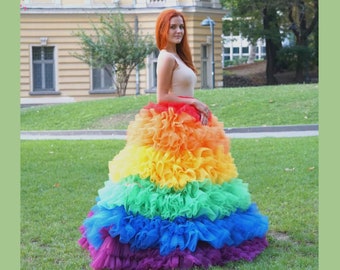 May include: A vibrant rainbow-colored dress with layers of ruffled fabric. The dress transitions through the colors of the rainbow, from red to purple. The dress is worn outdoors on a grassy lawn.