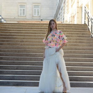 May include: A woman wearing a layered white tulle skirt and a top with neon pink, yellow, and black abstract designs. The top has ruffled sleeves. The woman is standing on a set of outdoor stairs.