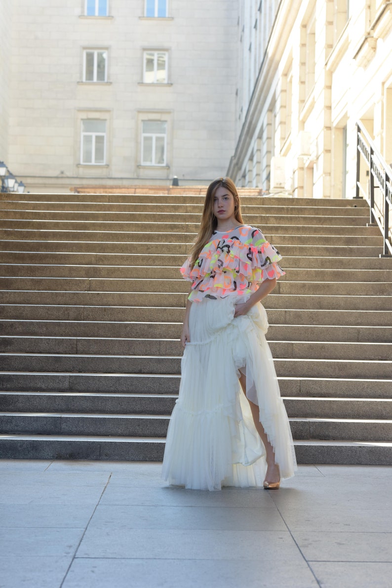 May include: A woman wearing a layered white tulle skirt and a top with neon pink, yellow, and black abstract designs. The top has ruffled sleeves. The woman is standing on a set of outdoor stairs.