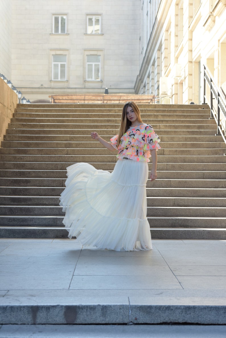 May include: A woman's outfit featuring a tiered white skirt and a ruffled top with pink, yellow, and black accents. The outfit is worn outdoors in front of a stone staircase and building.