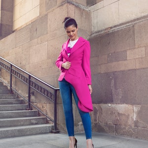 May include: A woman wearing a bright pink blazer with an asymmetrical design, paired with blue jeans and dark heels. The blazer has a unique cut and a long, flowing panel. The outfit is completed with a white t-shirt.