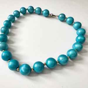 May include: A turquoise beaded necklace with gold-colored accents. The necklace features large, round beads in a vibrant turquoise hue, strung together with small gold-colored connectors and a clasp. The beads have a glossy finish.