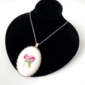 May include: Oval pendant necklace with a white background and a floral design featuring pink and purple flowers. The pendant is framed with a silver-colored metal and hangs from a delicate silver chain. The necklace is displayed on a black mannequin.