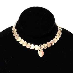 May include: A silver necklace with heart-shaped links. The necklace is displayed on a black velvet surface. A small heart-shaped charm hangs from the necklace.