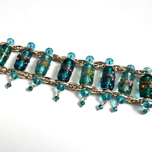 Op de afbeelding: Een goudkleurige kettingarmband met acht cilindervormige, blauwgroene glaskralen met bloemmotieven. De armband is versierd met kleine, bungelende lichtblauwe kristalkralen langs de randen en is afgewerkt met een staafsluiting.