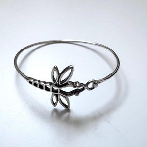 Pode incluir: Uma pulseira de metal em tom prateado com um motivo central de libélula. A libélula tem asas vazadas e um design de corpo segmentado. A pulseira é uma banda circular fina que se conecta ao ornamento da libélula em um dos lados.