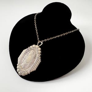 Op de afbeelding: Een zilverkleurige metalen medaillonhanger met een ingewikkeld gegraveerd patroon van bladmotieven en een centrale ovale plaat, hangend aan een bijpassende zilverkleurige schakelketting. Het medaillon heeft een geschulpte rand.