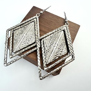 May include: A pair of silver-toned dangle earrings featuring a double diamond-shaped frame design. The outer frame has a textured edge, while the inner diamond plate displays an intricate embossed geometric pattern with Greek key-style motifs.